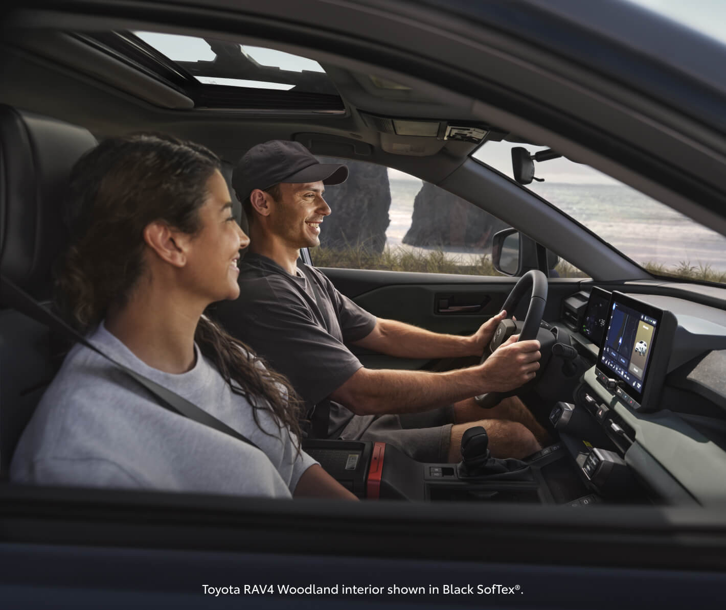 Side window view showcasing a couple driving a Toyota RAV4 Woodland Ed along a scenic coastal beach.