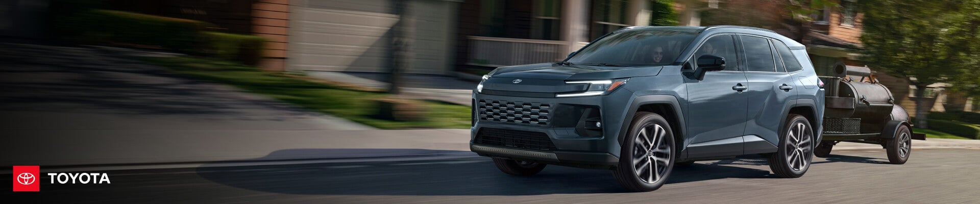 Three-quarter motion view of a 2026 Toyota RAV4 in Blueprint driving down a suburban street towing a trailer-mounted smoker barbecue.