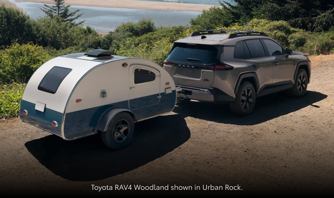 Rear high-angle view of a 2026 Toyota RAV4 Woodland Edition in Urban Rock towing a vintage-style teardrop camper along a dirt road.
