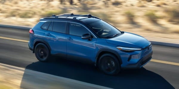 Blue Toyota Corolla Cross Hybrid driving on a road, shown from an elevated front three-quarter view.